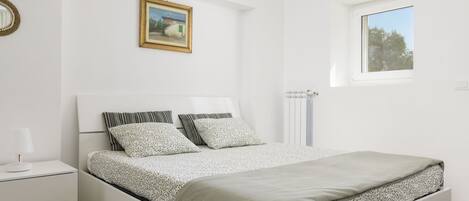 Ein Schlafzimmer mit einem Bett, einem Nachttisch, einem Heizkörper, einem Fenster und einem Wandbild.
