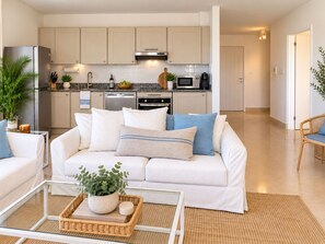 Una sala de estar moderna con un sofá blanco, una mesa de centro de vidrio y una cocina al fondo.