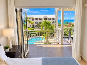 A hotel room with a bed, a nightstand, a lamp, and a view of a pool and resort buildings. - Sandbox Villa Stylish 2-bedroom on West Coast in  St. James - steps to beach (Weston)