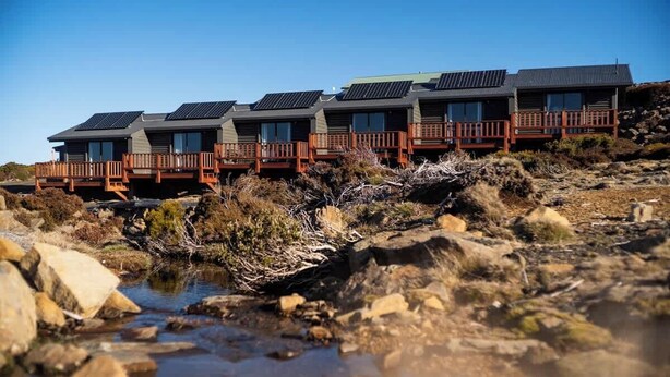 Une rangée d’immeubles modernes à plusieurs étages, dotés de panneaux solaires sur les toits, est construite sur un terrain rocheux, avec un ruisseau en premier plan.