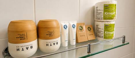 Un estante de baño con varios artículos de aseo, incluyendo un recipiente de bambú, un tubo, un rollo de pañuelos de papel y una pequeña bolsa marrón.