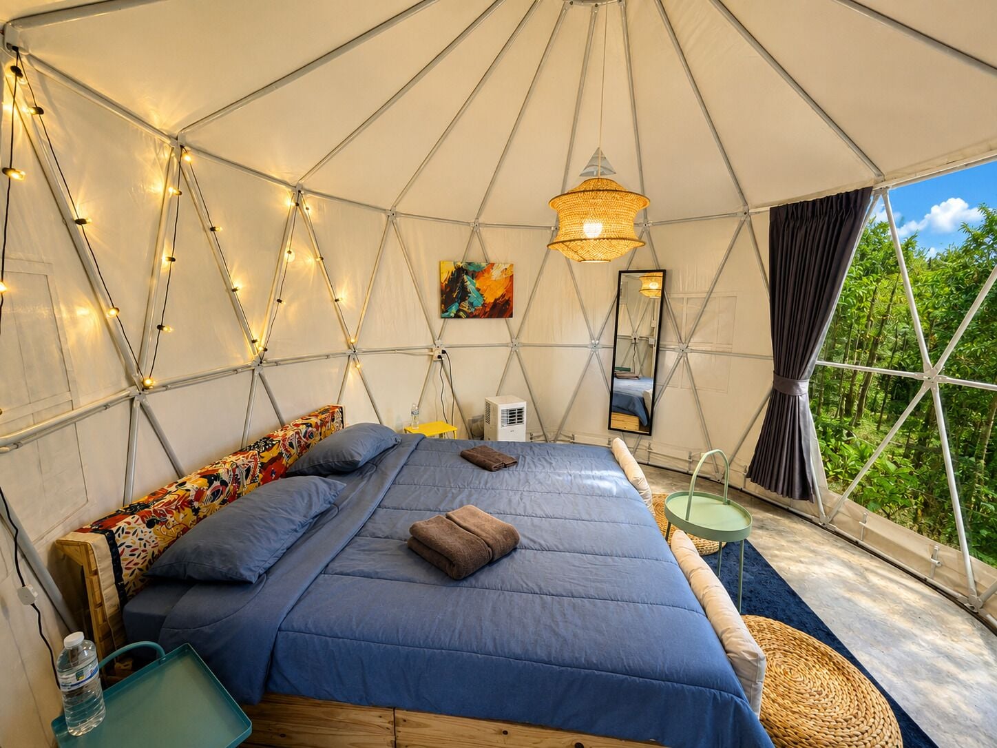 A tented accommodation room with a bed, pillows, a hanging light fixture, a mirror, and a view of greenery outside.