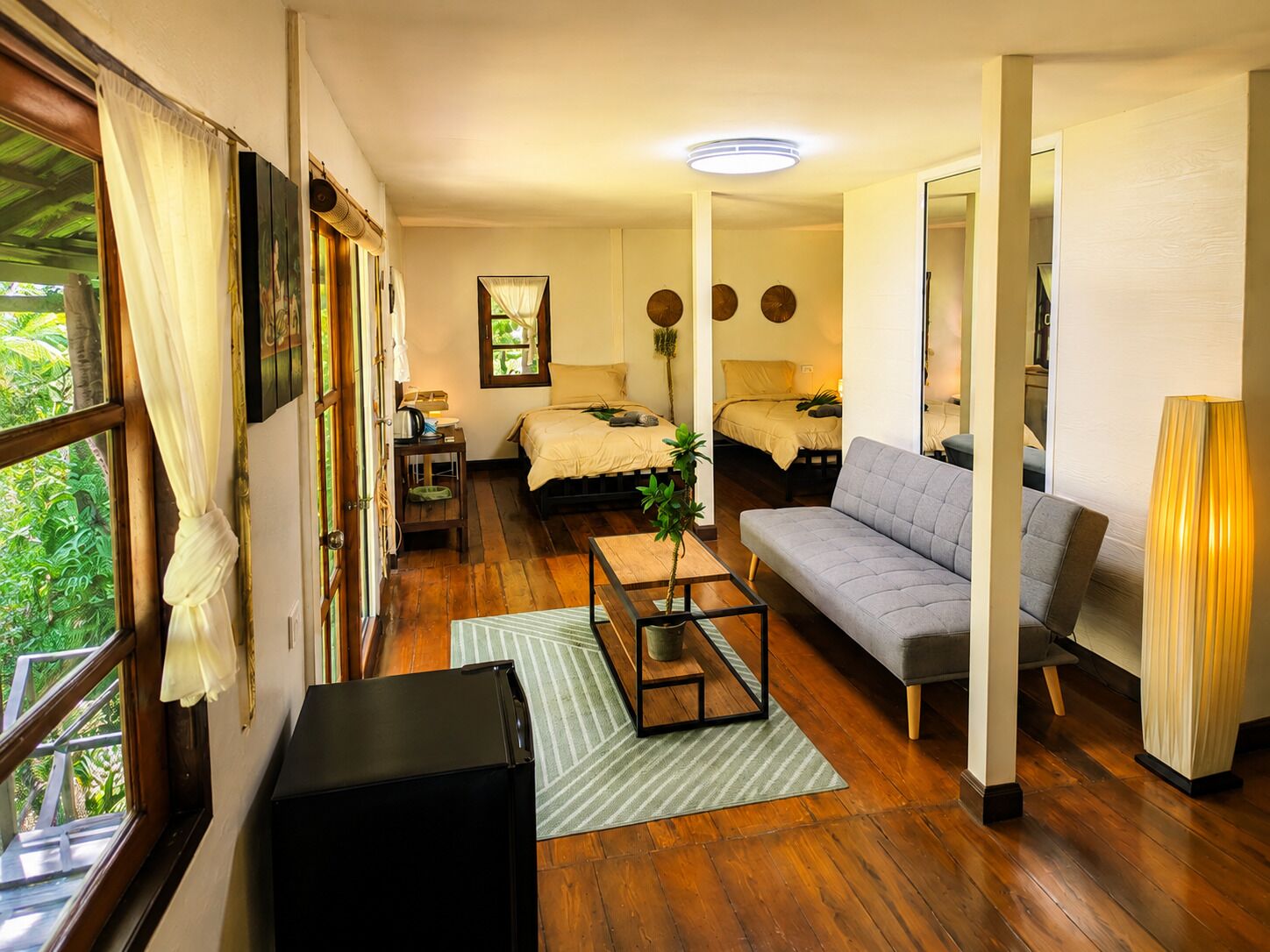A room with a bed, a sofa, a coffee table, and a lamp. There is a window with curtains, a door, and a mirror.