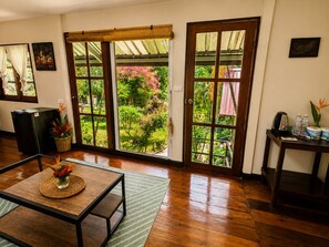 Een kamer met houten vloer, een tafel met bloevas, een koelkast, een raam met uitzicht op groen en een deur met uitzicht op een tuin.