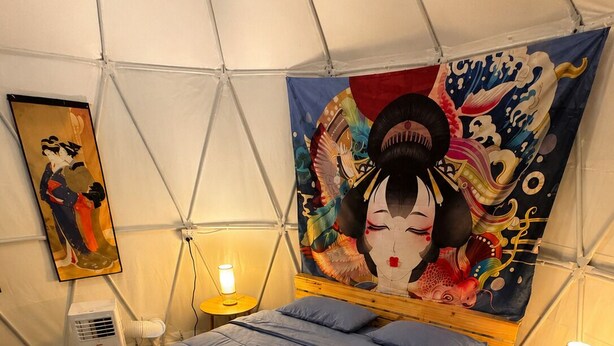 Une pièce comprenant un lit, une chaise, une petite table, une tapisserie murale et un climatiseur fixé au mur.