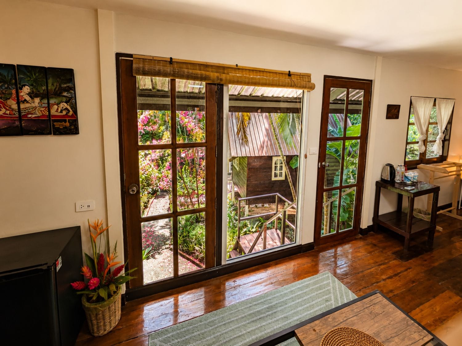 Una habitación con piso de madera, un televisor, una mesa con lámpara y vistas a un jardín por un ventanal.