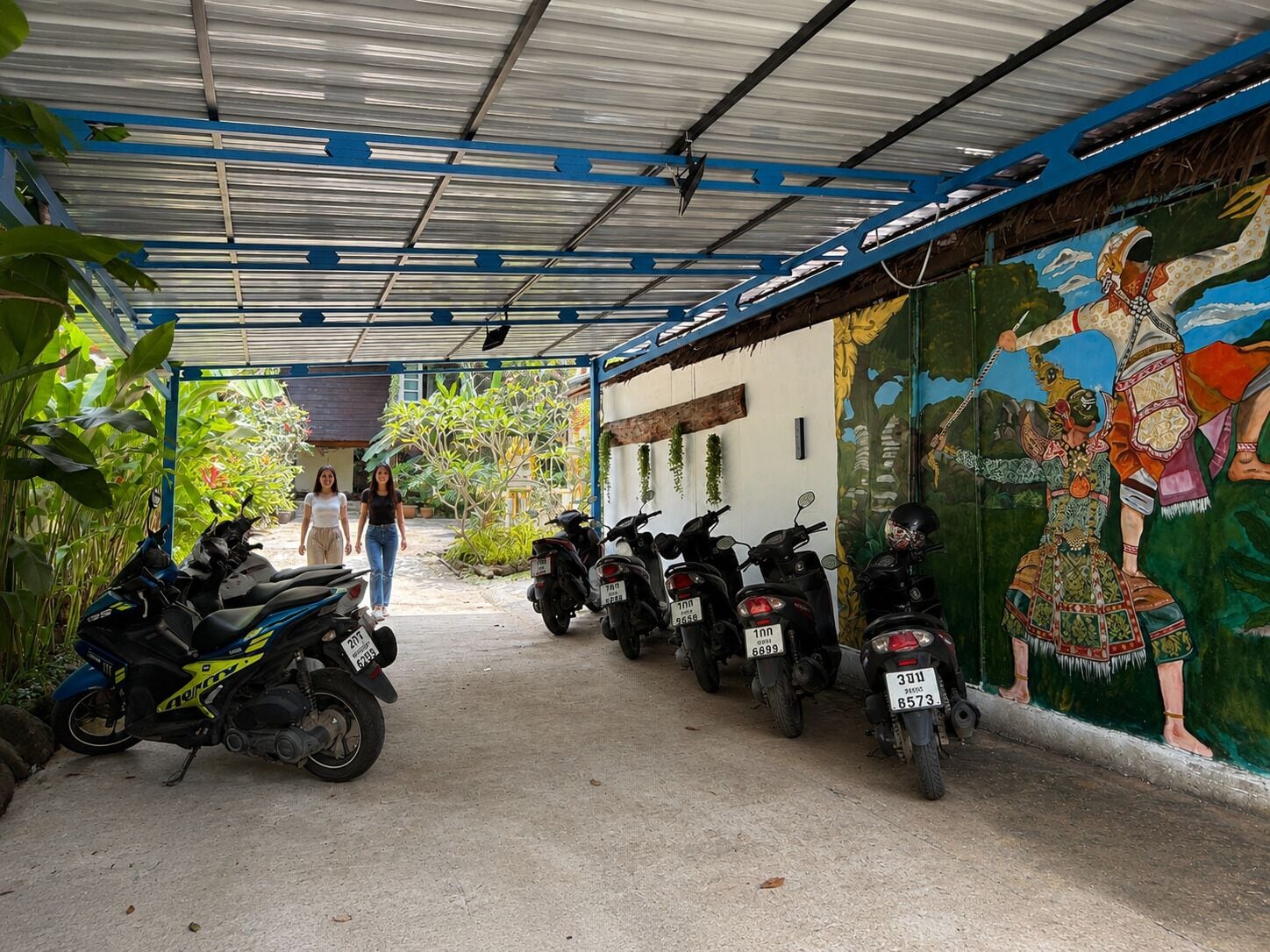 A covered parking area with motorcycles parked, a mural on the wall, and a person walking in the distance.