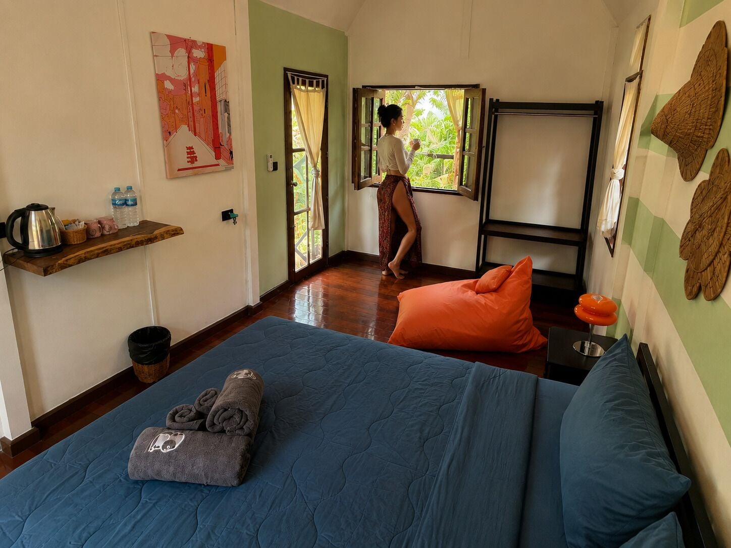 A room with a bed, a window with curtains, a shelf with items, and a wall with artwork.