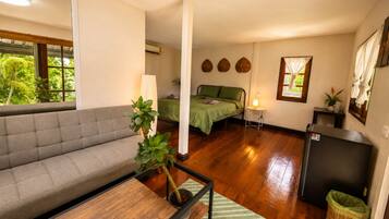 Ein Zimmer mit einer Couch, einem Bett, einem kleinen Tisch, einer Lampe, einem Kühlschrank und einem Fenster mit Vorhängen.