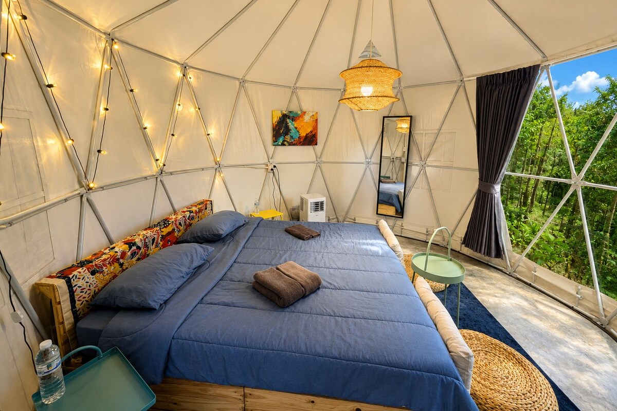 A tented accommodation room with a bed, pillows, a hanging light fixture, a mirror, and a view of greenery outside.