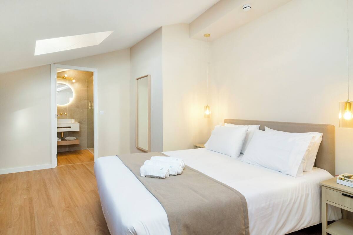 Une chambre d’hôtel moderne avec un grand lit, une table de chevet, une commode, un miroir et une fenêtre de toit.