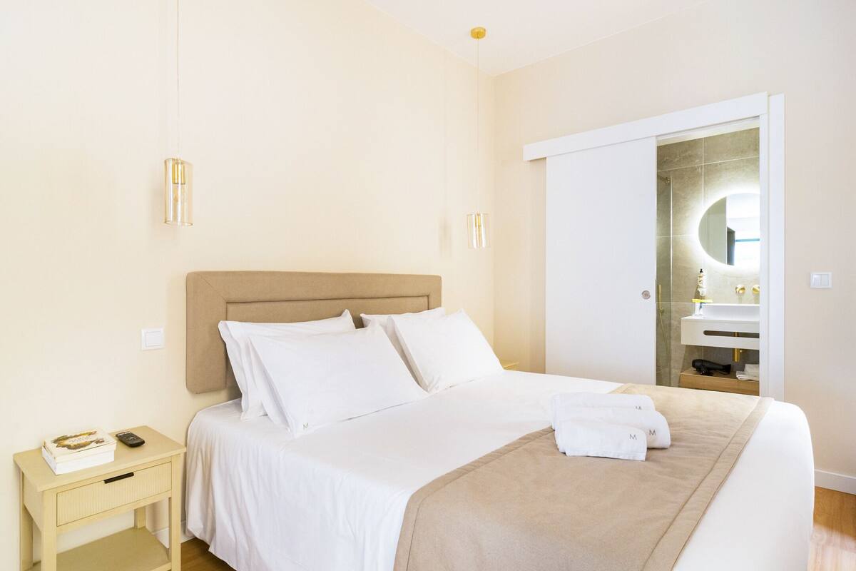 Un lit bien fait, avec du linge de lit blanc, une table de chevet sur laquelle se trouvent un livre et une télécommande, et une salle de bain avec un miroir et un lavabo.