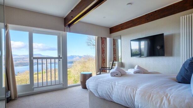 A bedroom with a large bed, a TV mounted on the wall, and a view of a lake through a sliding door. - Paddock House (Windermere)