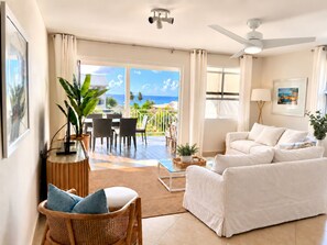 A modern living room with a white sofa, a dining area with chairs, a ceiling fan, and a view of the ocean. - Sandbox Villa Stylish 3-bedroom on West Coast in  St. James - steps to beach (Weston)