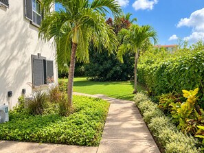 A pathway leading to a building with a white exterior and shutters, surrounded by lush greenery and a well-maintained lawn.