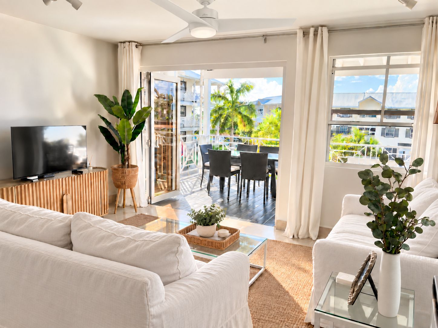 A modern living room with a flat-screen TV, white sofas, a glass coffee table, and a balcony with outdoor furniture.