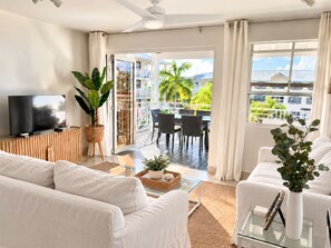 A modern living room with a flat-screen TV, white sofas, a glass coffee table, and a balcony with outdoor furniture.