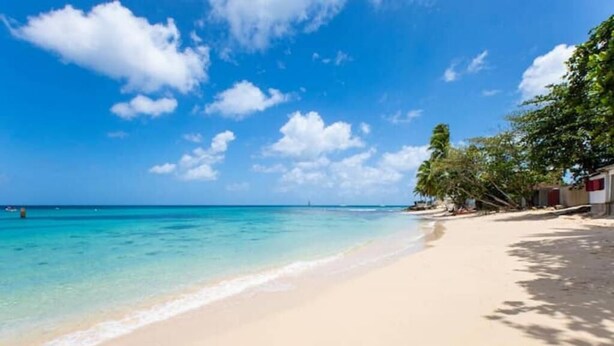 Une plage à l’eau turquoise et limpide, un rivage sablonneux et une rangée de bâtiments aux toits rouges.