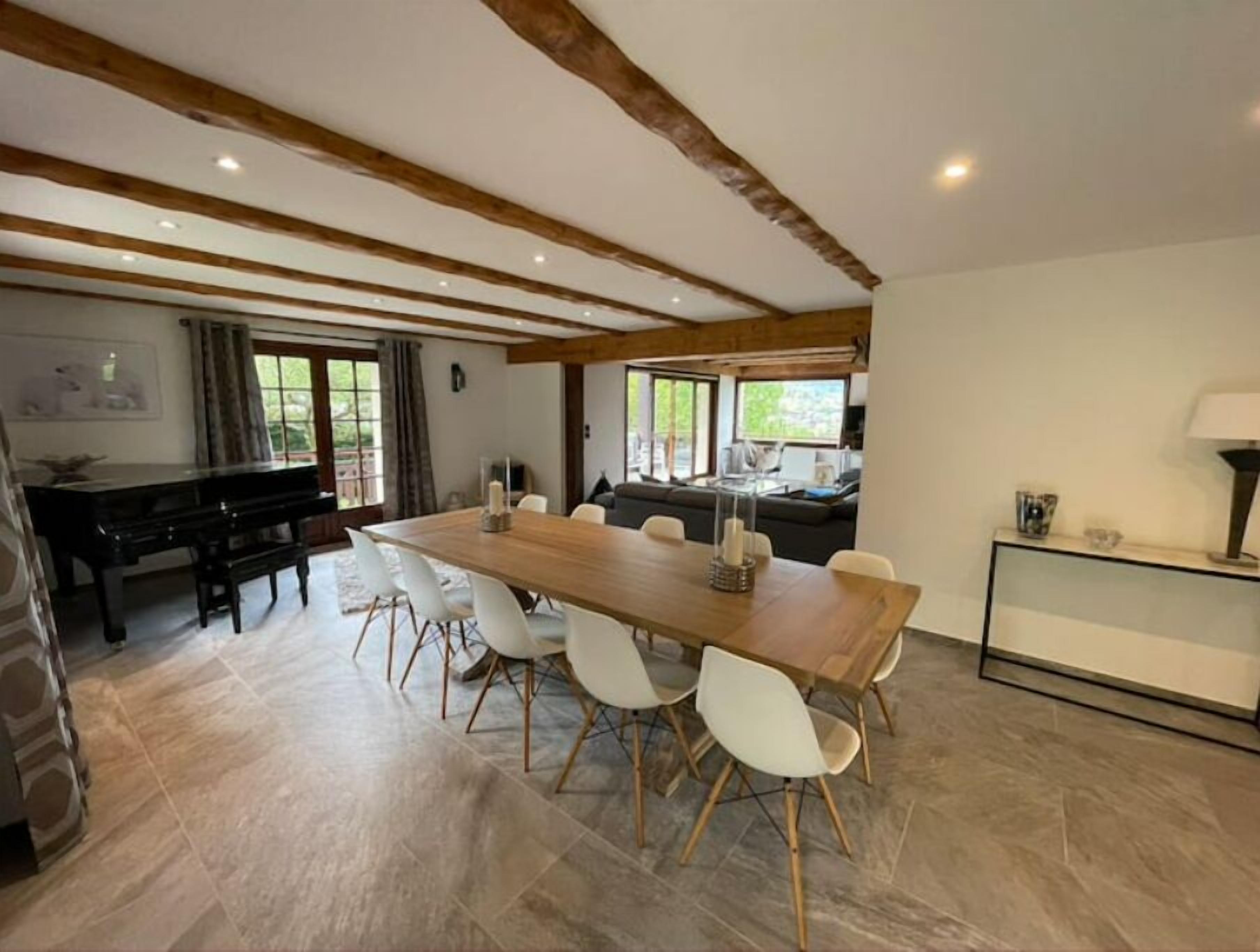 A spacious dining area with a long wooden table and white chairs, a piano, and a sofa in the background.