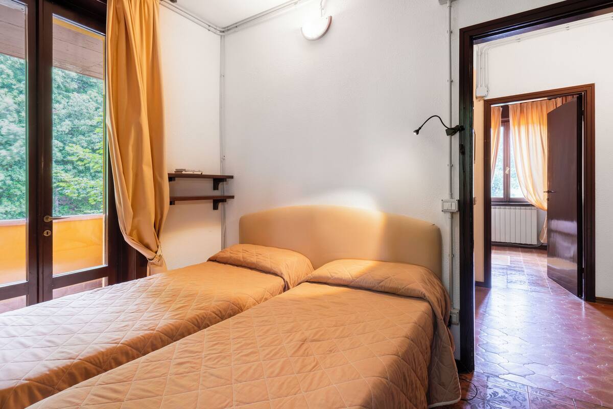 Una habitación con dos camas, una ventana con cortinas, una lámpara fijada a la pared y una puerta que da a otra habitación.