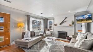 A modern living room with a fireplace, a sofa, a coffee table, and a TV mounted on the wall. - Historic Mountain Ave Bungalow – Walk Everywhere! (Fort Collins)