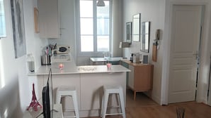 Een moderne keuken met witte kasten, een eethoek met een witte tafel en twee krukken, een groot raam met gordijnen, en een deur die leidt naar een andere kamer.