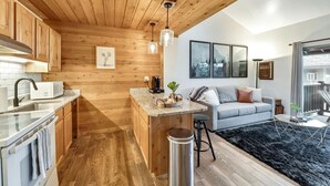 Een moderne keuken met houten kastjes en een centraal eiland, een woonruimte met een bank en salontafel, en een groot raam met uitzicht.
