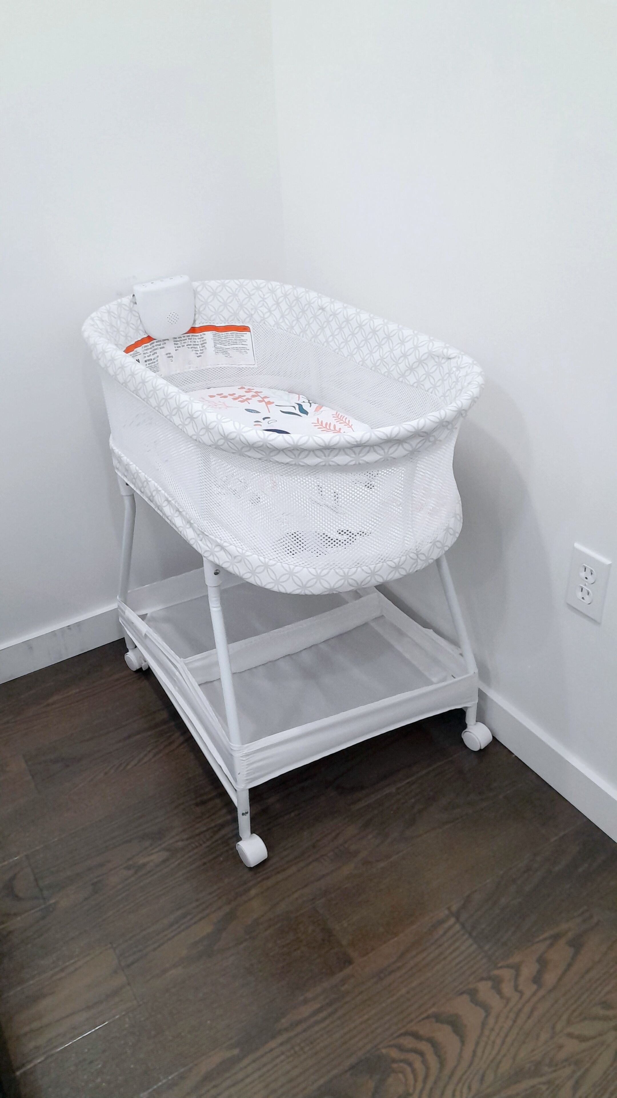 A white baby bassinet with a mesh top and a lower shelf, placed against a white wall with a wooden floor.