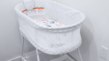 A white baby bassinet with a mesh top and a lower shelf, placed against a white wall with a wooden floor.