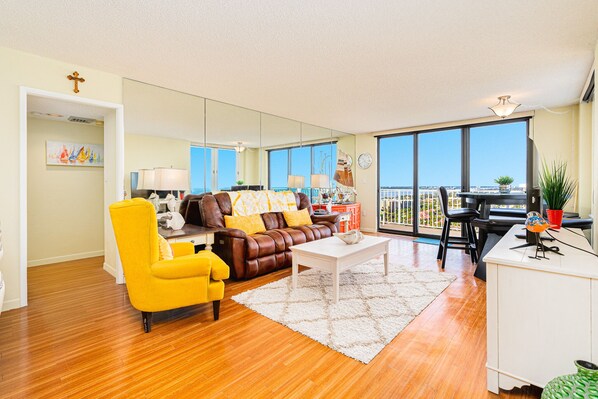 A spacious living room with a brown leather sofa, yellow armchair, and white coffee table. A dining area with a table and chairs is visible through large windows. - Oceanfront Penthouse-Heated Pool-Oceans 3 #2206 (Daytona Beach Shores)