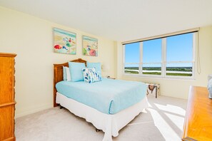 A bedroom with a large window, a bed with blue bedding, and two wall-mounted paintings. - Oceanfront Penthouse-Heated Pool-Oceans 3 #2206 (Daytona Beach Shores)