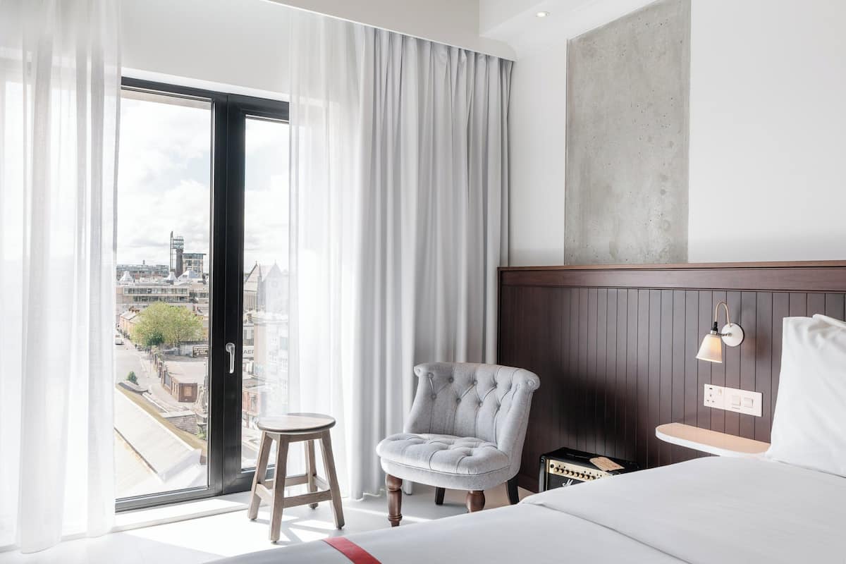 A hotel room with a large window, a bed, a chair, a small table, and a wall-mounted light fixture.