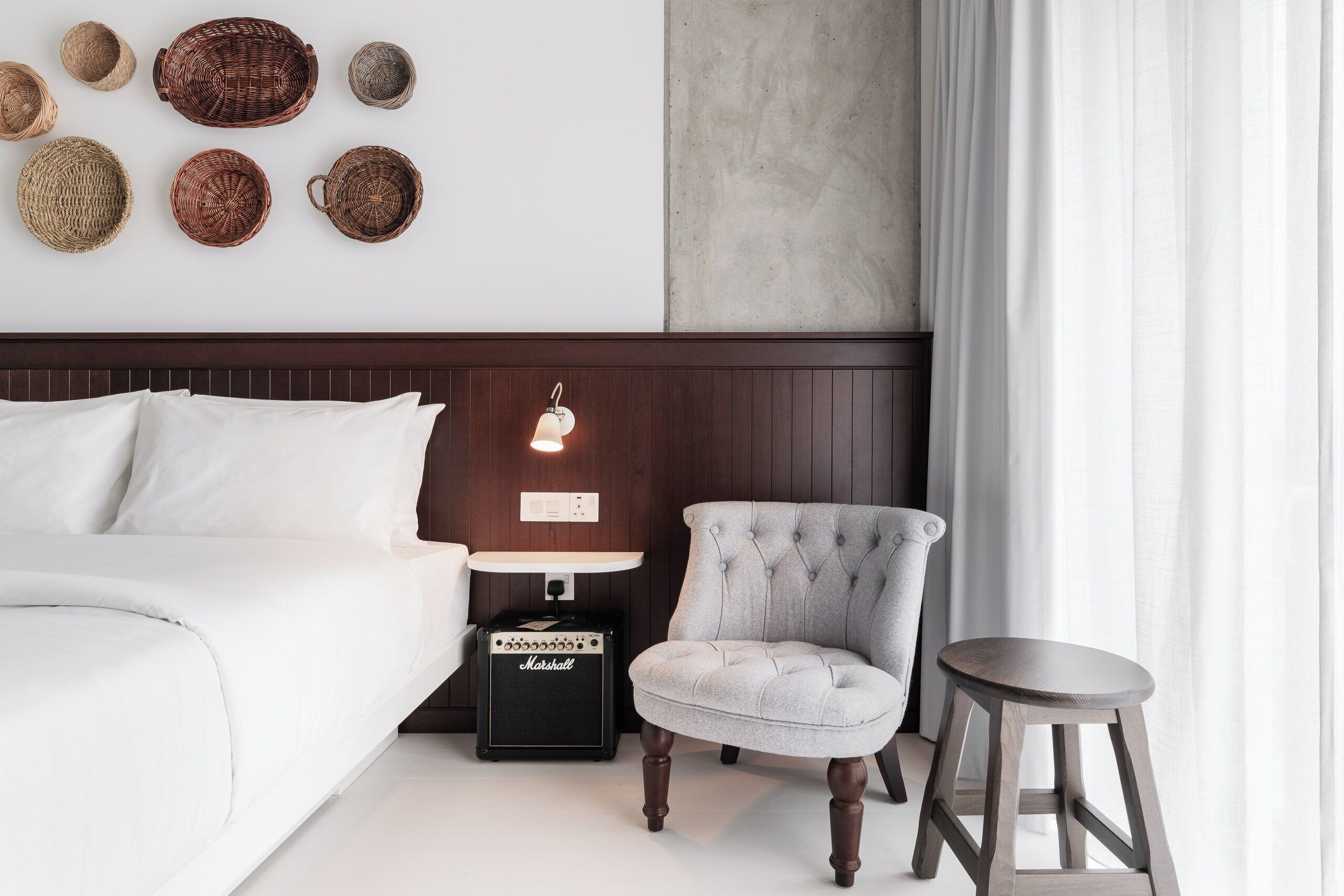 A neatly arranged bedroom with a bed, a chair, a stool, a nightstand with a lamp, and a wall-mounted basket collection.
