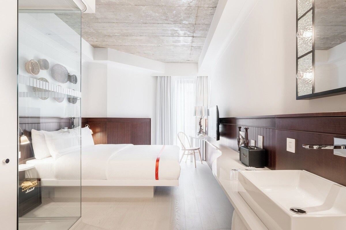 A modern hotel room with a glass partition, a bed, a bathroom with a large sink, and a wooden headboard.