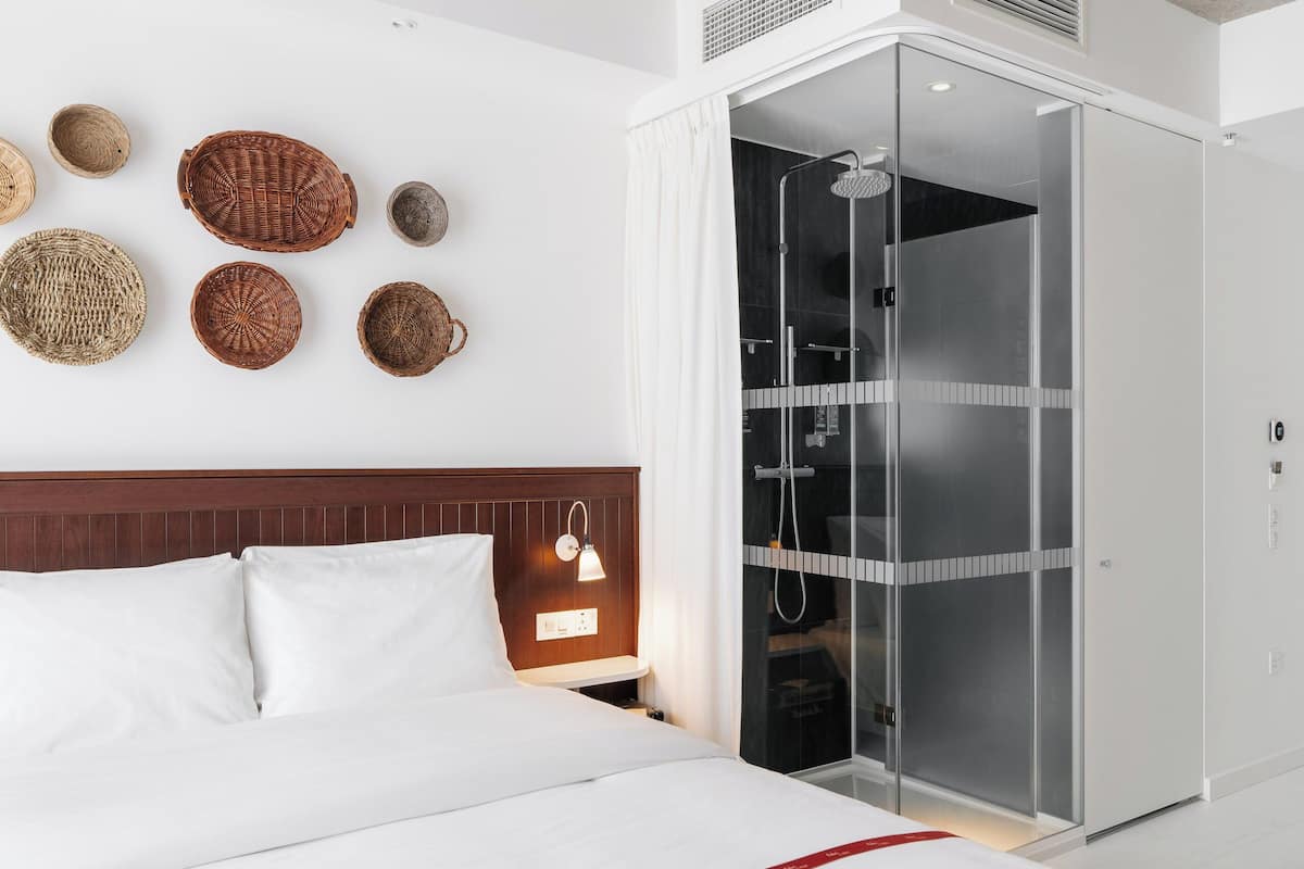 A hotel room with a bed, a nightstand, a wall-mounted lamp, a shower enclosure, and several woven baskets on the wall.