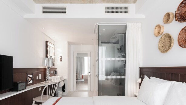 A hotel room with a bed, a desk with a chair, a television, a mirror, and a bathroom with a shower and toilet.
