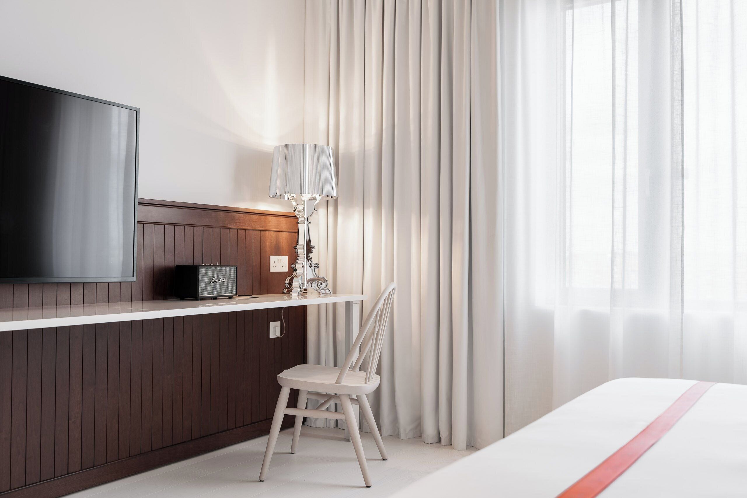 A hotel room with a flat-screen TV, a bedside table with a lamp, a chair, and a bed with a red stripe.