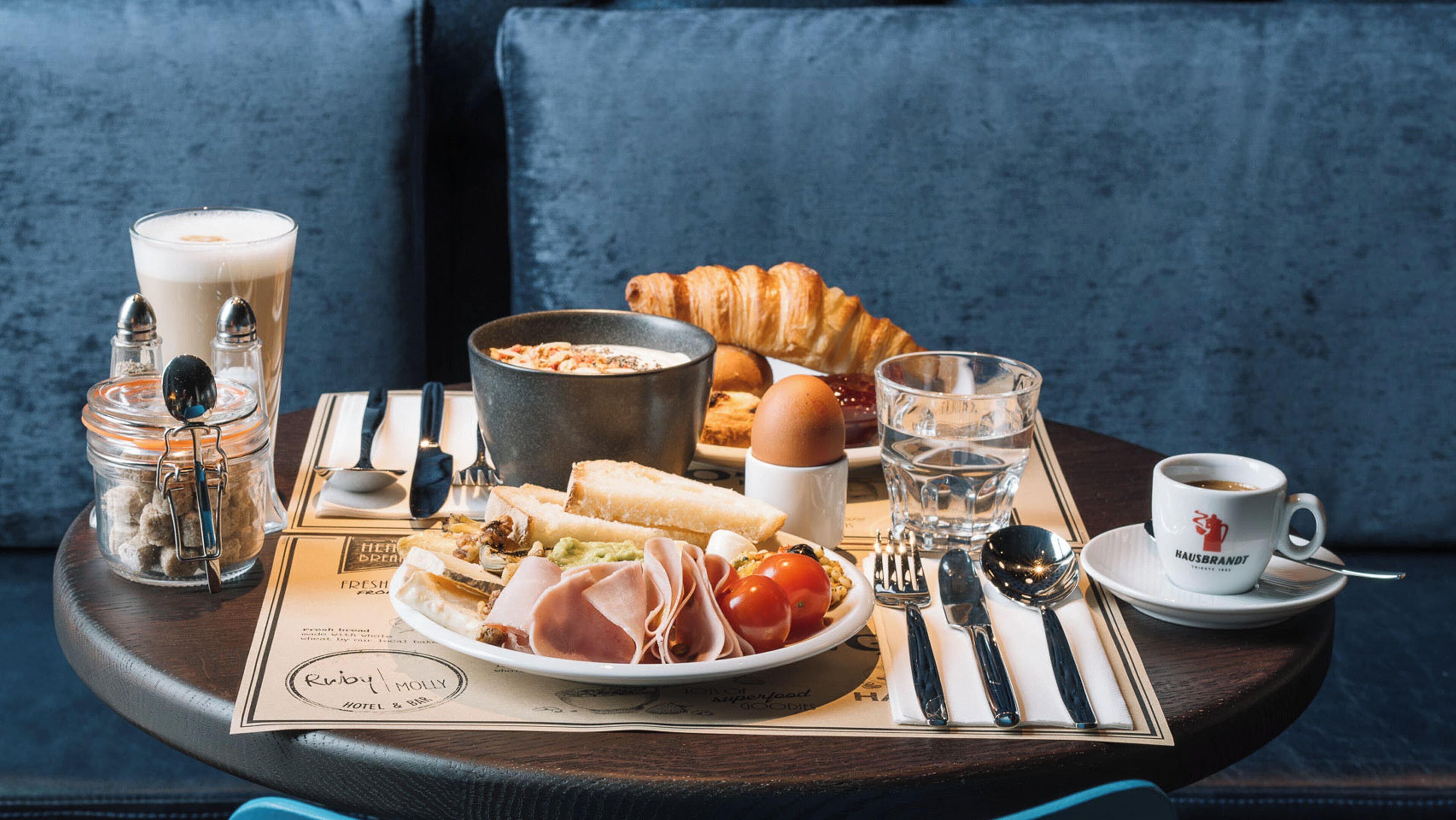 A breakfast tray with croissants, eggs, tomatoes, ham, and a bowl of soup on a table with a blue cushion in the background.