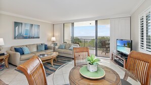 A spacious living room with a large window, a flat-screen TV, a sofa, and a dining area with chairs and a table.