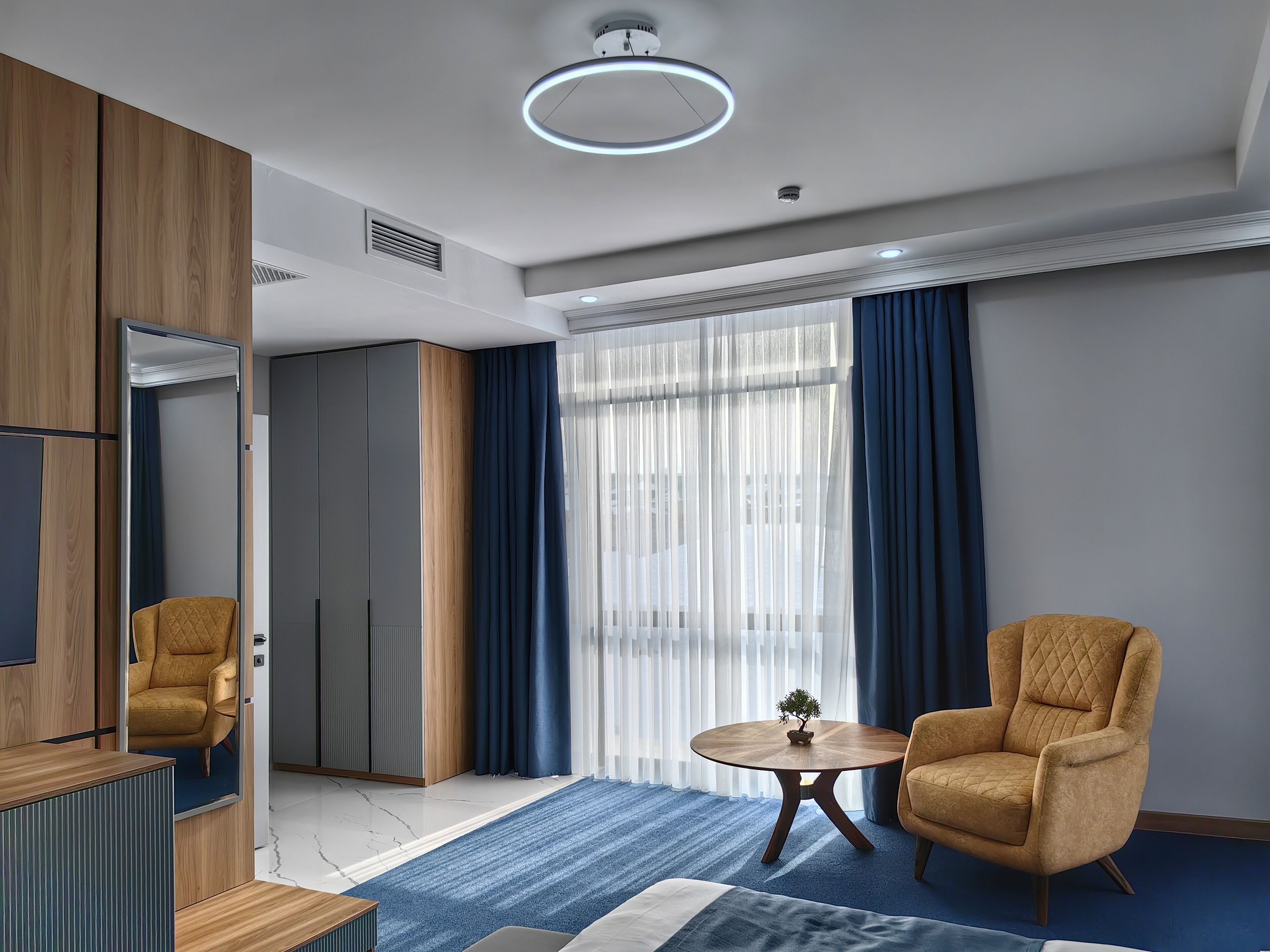 A modern hotel room with a large bed, a round wooden table, a comfortable armchair, a mirror, and a wardrobe.