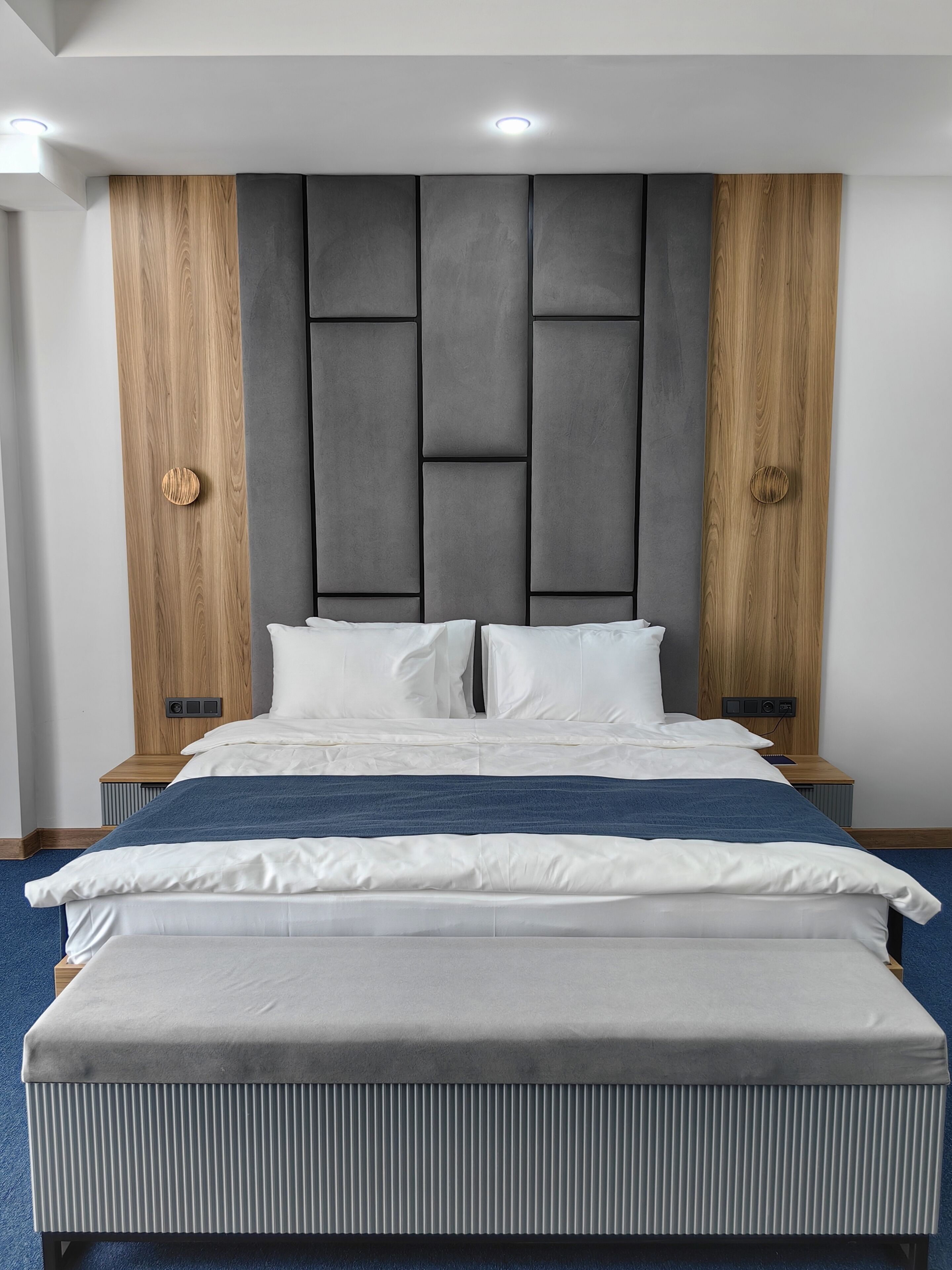 A modern hotel room with a large bed, a padded headboard, and wooden paneling on the walls.