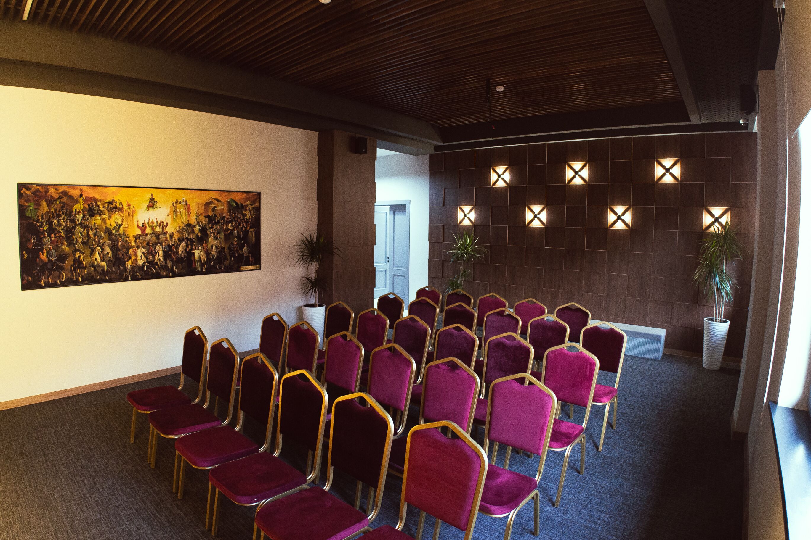 Ein Konferenzraum mit Reihen lila Stühlen, einem großen Gemälde an der Wand und einer Holzbalkendecke.