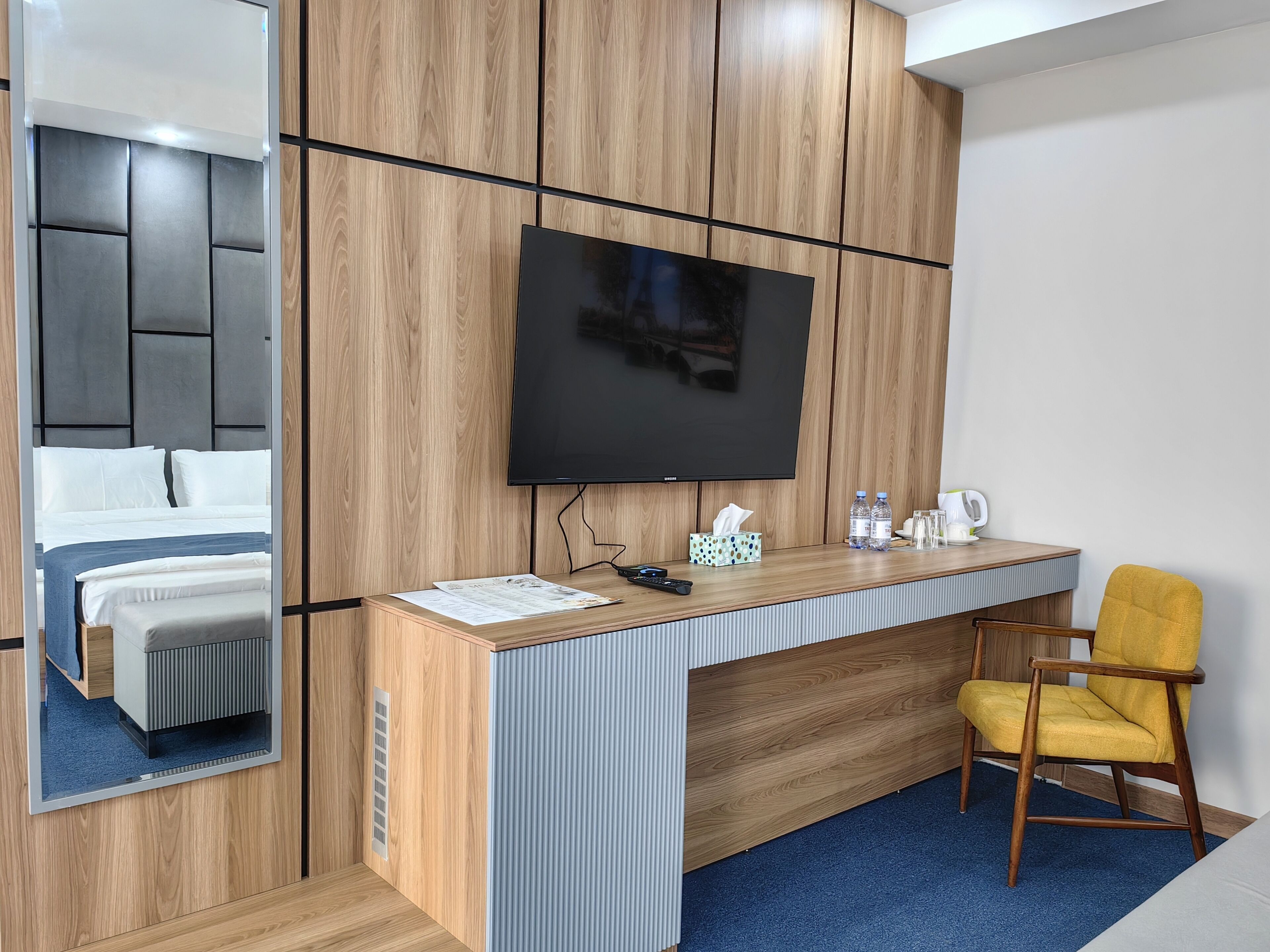 A hotel room with a wooden desk, a flat-screen TV, a yellow chair, and a bed with white bedding.