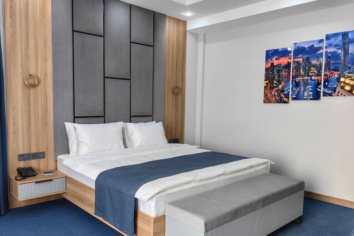 Una habitación de hotel moderna con una cama grande, una mesita de noche con teléfono y libreta, una obra de arte en la pared que representa un paisaje urbano, y un banco empotrado al pie de la cama.