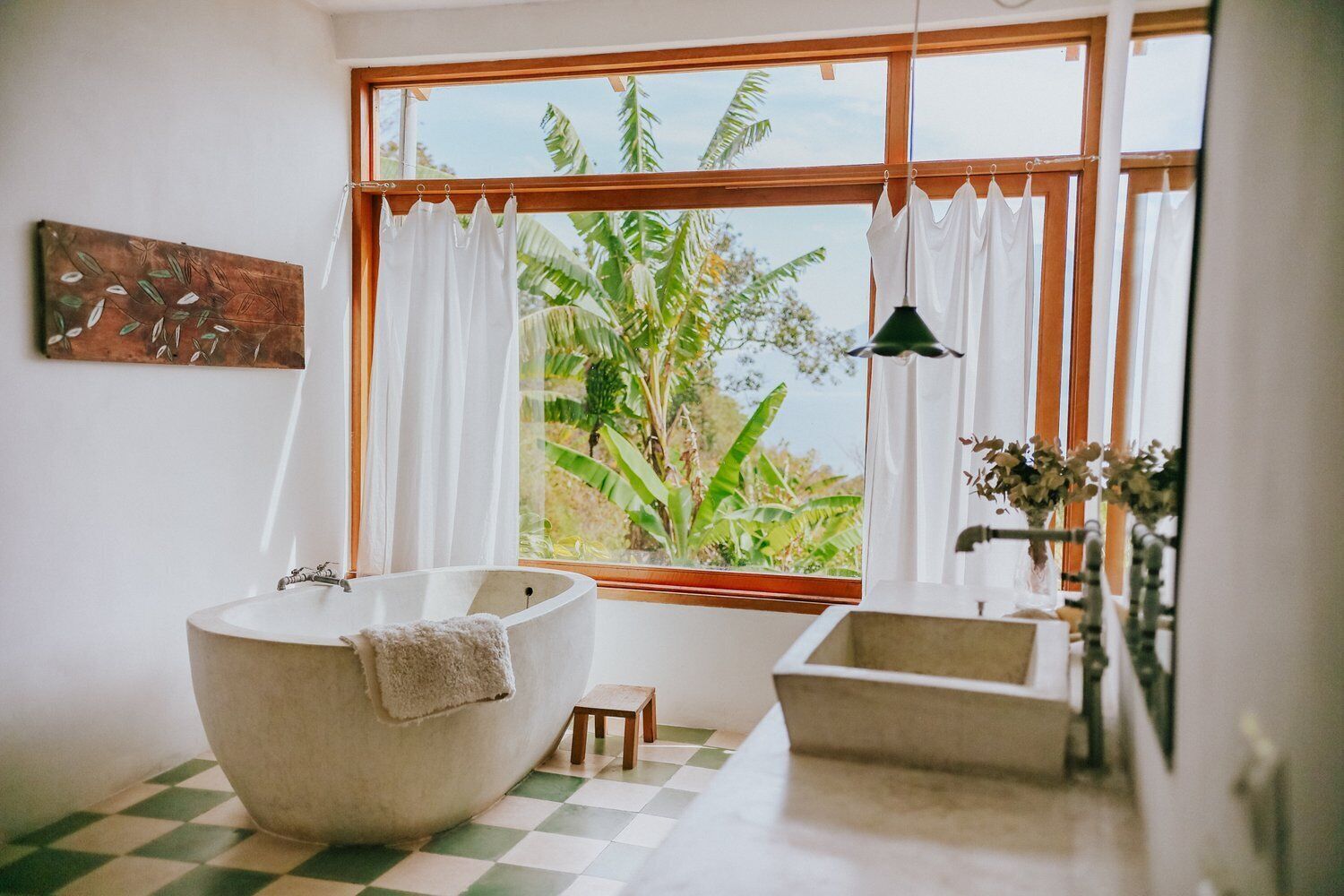 Un baño con una bañera independiente, un taburete de madera pequeño y una ventana con cortinas.