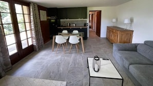 Ein modernes Wohnzimmer mit grauem Sofa, einem Couchtisch und einem Essbereich mit Tisch und Stühlen. Im Hintergrund ist eine Küche mit Blick ins Freie zu sehen.
