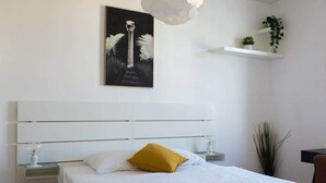 Een slaapkamer met een wit bed, een geel kussen, een schilderij van een vogel, een witte plank met planten en een nachtkastje met een lamp.