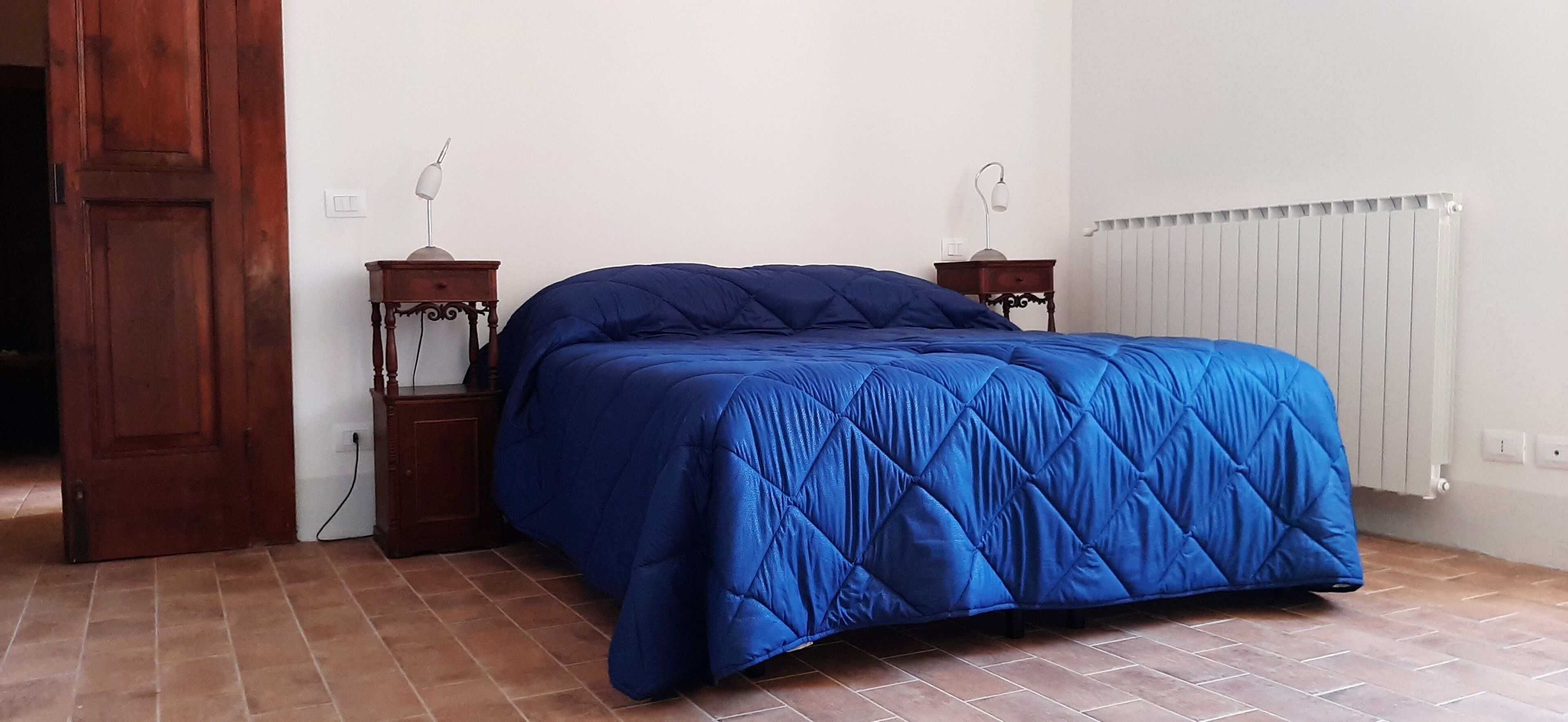 Een slaapkamer met een groot bed, twee nachtkastjes, een houten deur, een radiator en een betegelde vloer.