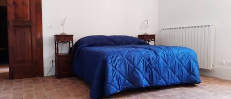 A bedroom with a large bed, two bedside tables, a wooden door, a radiator, and a tiled floor.