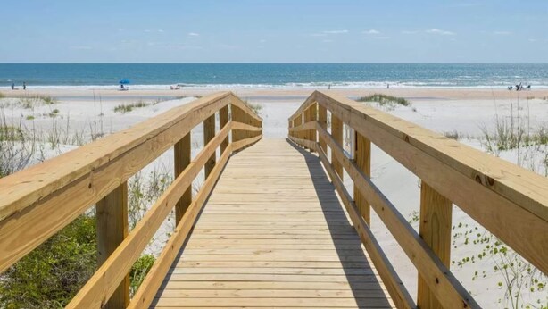 Un muelle de madera que lleva a una playa con arena y vegetación escasa.